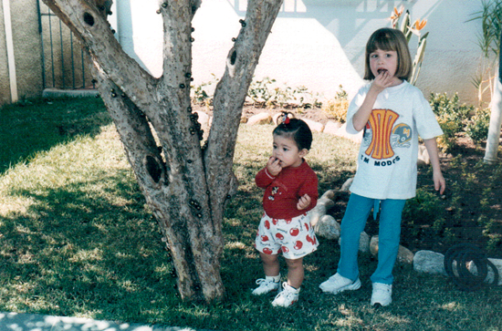 Duas crianças em duas idades diferentes embaixo de uma jabuticabeira, comendo jabuticabas. Na esquerda a Maria, com aproximadamente 2 anos, de camiseta vermelha e shorts com detalhes em vermelho, na esquerda Camila, aproximadamente 7 anos, de camisa branca e calça azul.